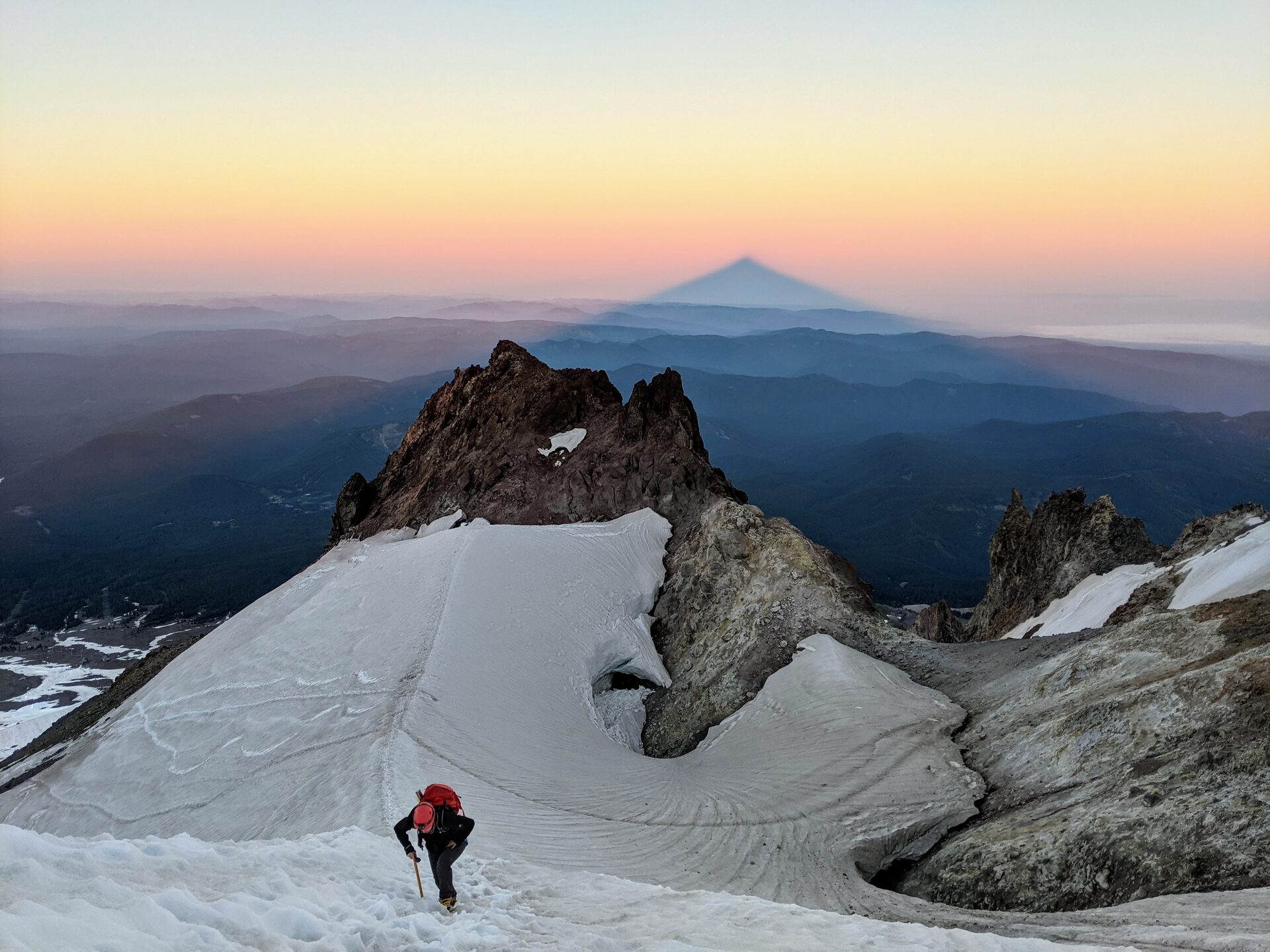 Dawn from high on Mt. Hood: a climber ascends a snowfield while the mountain casts its own triangular shadow onto the atmosphere.