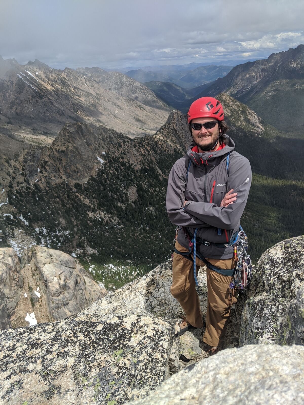 Adam stands on a granite summit block in a red helmet with a rack of climbing gear on his harness, arms crossed, ridgelines and a deep forested valley behind him.