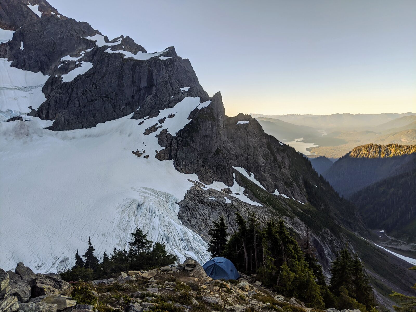 A blue tent pitched on a rocky bench beneath a glacier, with a lake and forested valley far below at sunrise.