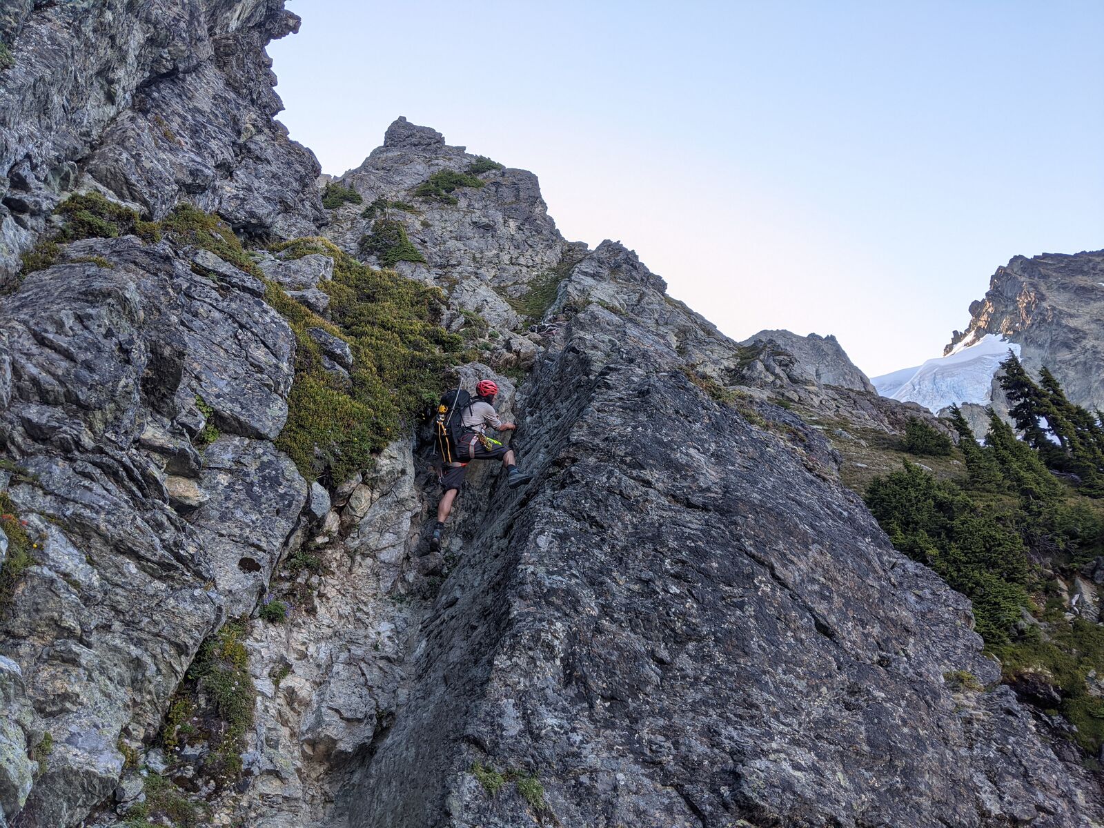 A climber scrambles up a rocky ridge step with an ice axe strapped to his pack, a glacier hanging in the basin behind.