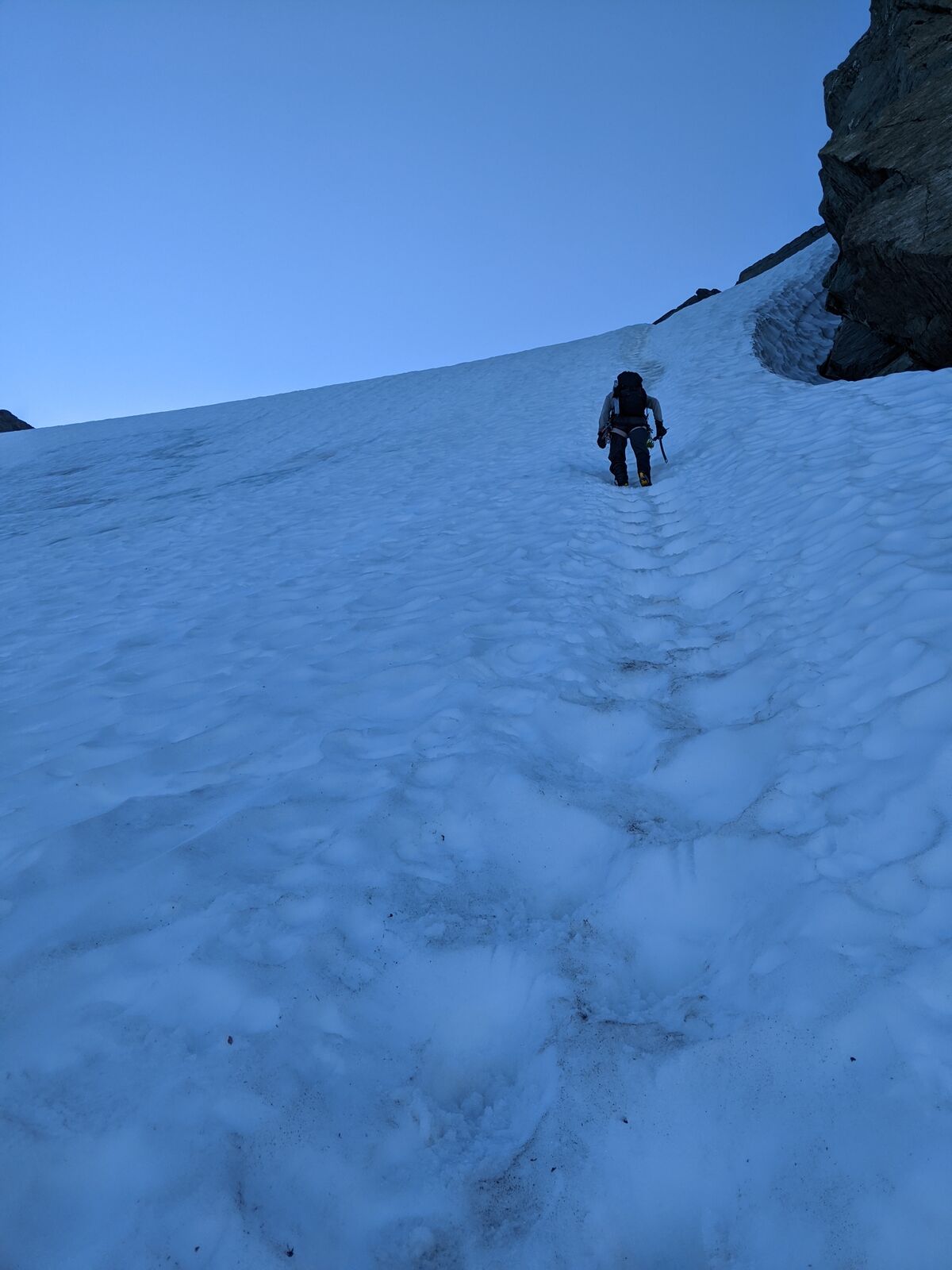 A climber ascending a steep snow couloir in blue pre-sun light, kicked steps trailing below.