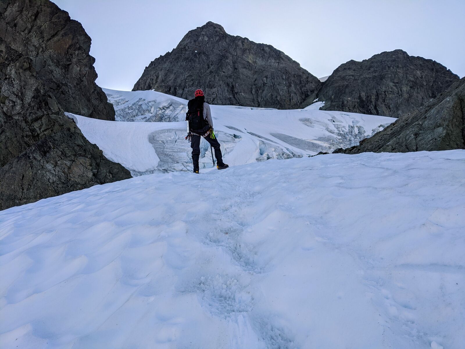 A climber in a red helmet stands on glacier ice below dark rock towers, studying the crevassed slope ahead.