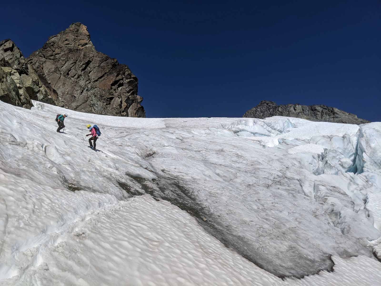 Two climbers descending a sun-softened glacier under deep blue sky, crevasse field opening to their right.