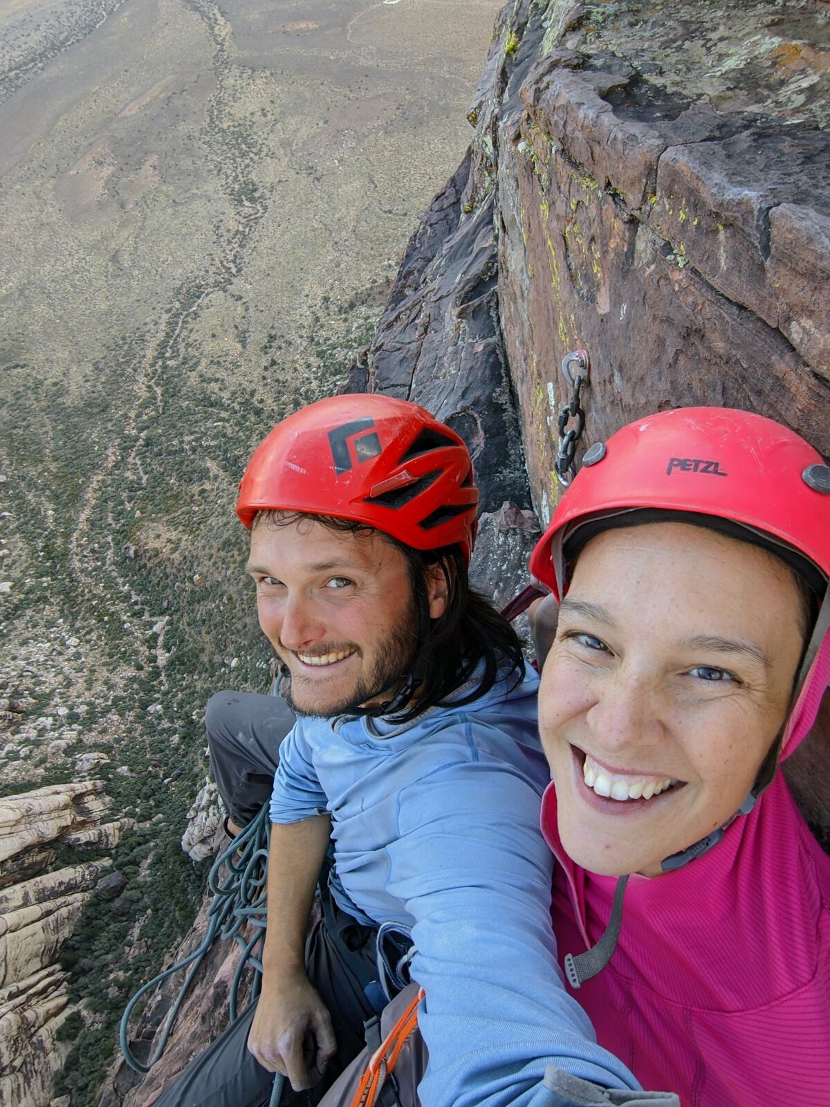 Two climbers grin from a small belay ledge hundreds of feet up a desert sandstone wall, the canyon floor far below.