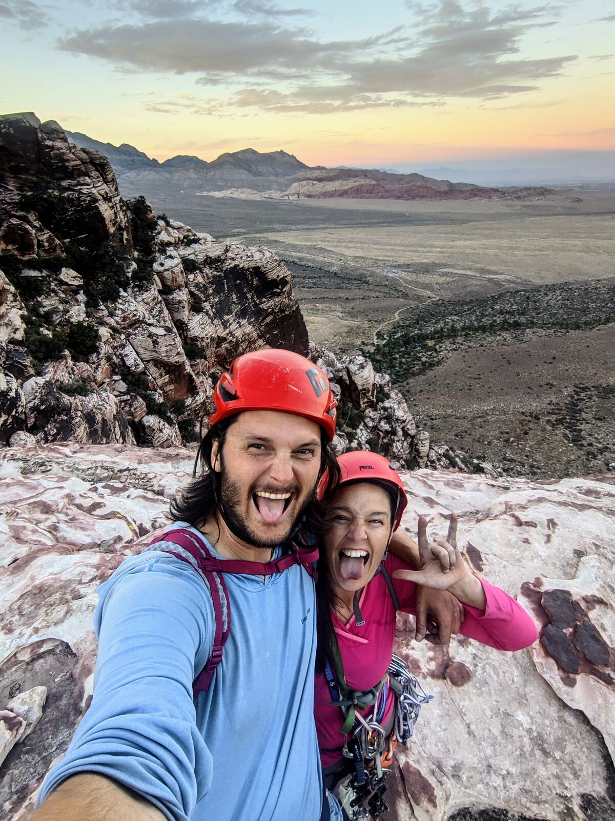 Two climbers in red helmets mug for a summit selfie at sunset, tongues out, desert canyon country glowing pink and gold behind them.