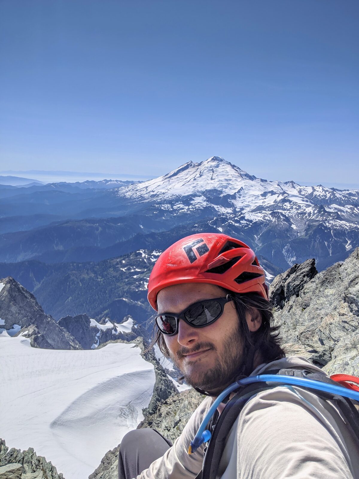 Adam W. Brown in a red climbing helmet and sunglasses on a rocky summit, a huge glaciated volcano rising behind him.